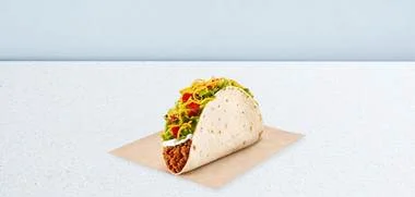 Soft taco filled with lettuce, tomato, cheese, and meat on a parchment square on a light surface.